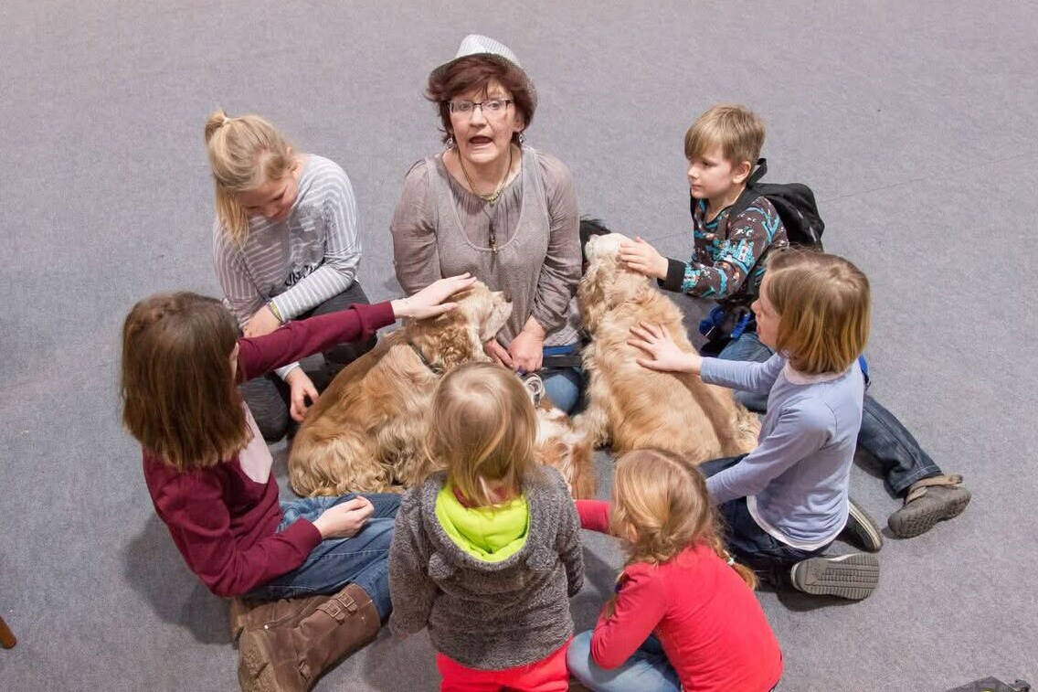 Eine Frau sitzt mit sechs Kindern im Kreis, in der Mitte liegen zwei Hunde, die gestreichelt werden